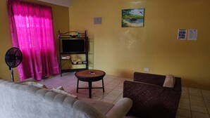 Une pièce comprenant un canapé, une petite table, un ventilateur, un téléviseur posé sur un meuble et un tableau accroché au mur.