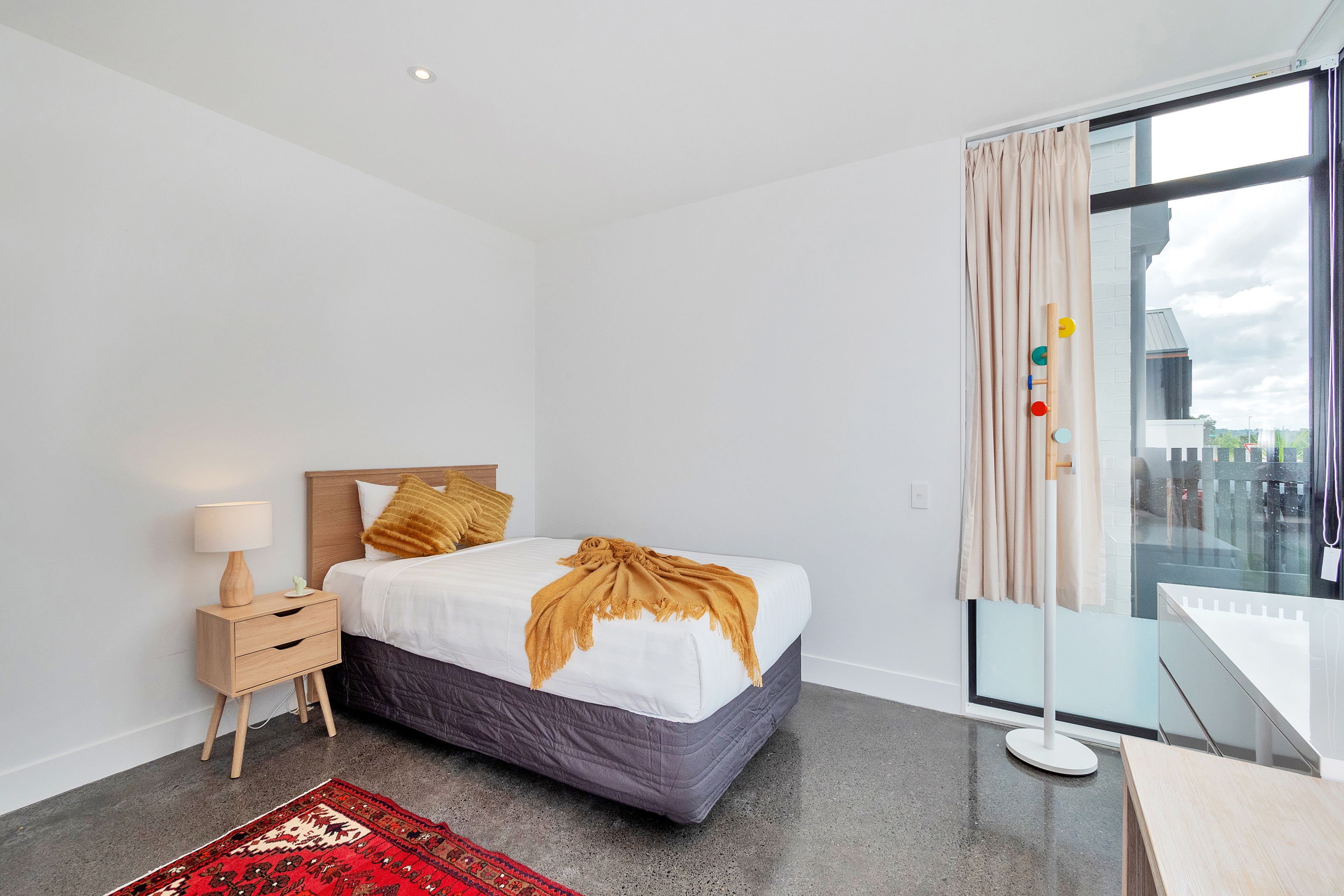 A modern hotel room with a large bed, a bedside table with a lamp, a red patterned rug, and a view of the city through a large window.