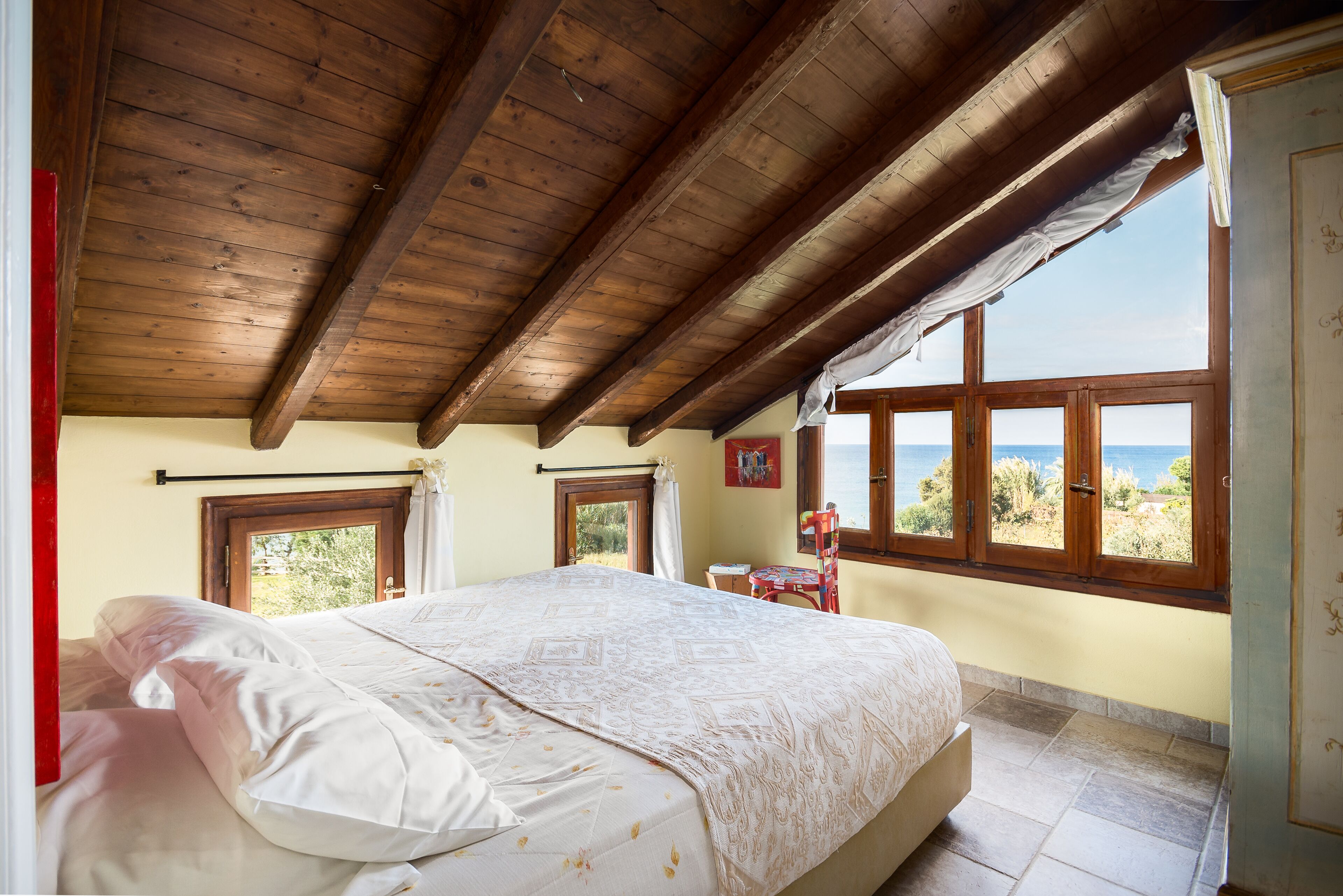 A bedroom with a wooden ceiling, a bed with white linens, a view of the sea through windows, and a small table with a chair.