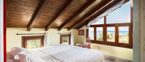 A bedroom with a wooden ceiling, a bed with white linens, a view of the sea through windows, and a small table with a chair.
