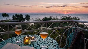 A table with two glasses of beer, a candle, and an open book, overlooking a sunset over the sea.