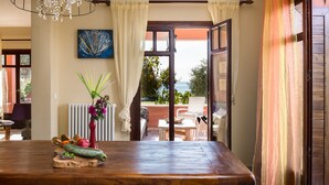 A wooden table with a fruit and vegetable centerpiece, a vase with flowers, and a view of an outdoor patio with a chair and table.