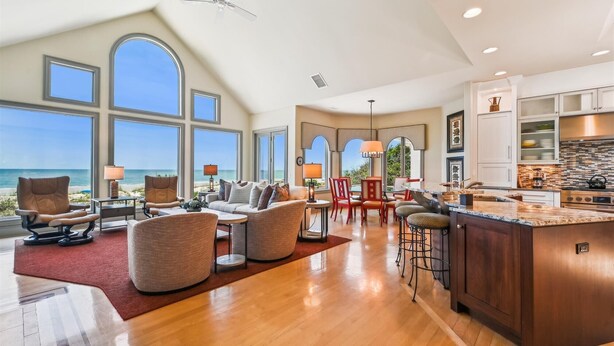 Een ruime woonkamer met grote ramen met uitzicht op de oceaan, een moderne keuken met een groot schiereiland en een gezellige zithoek met meerdere stoelen en een bank.