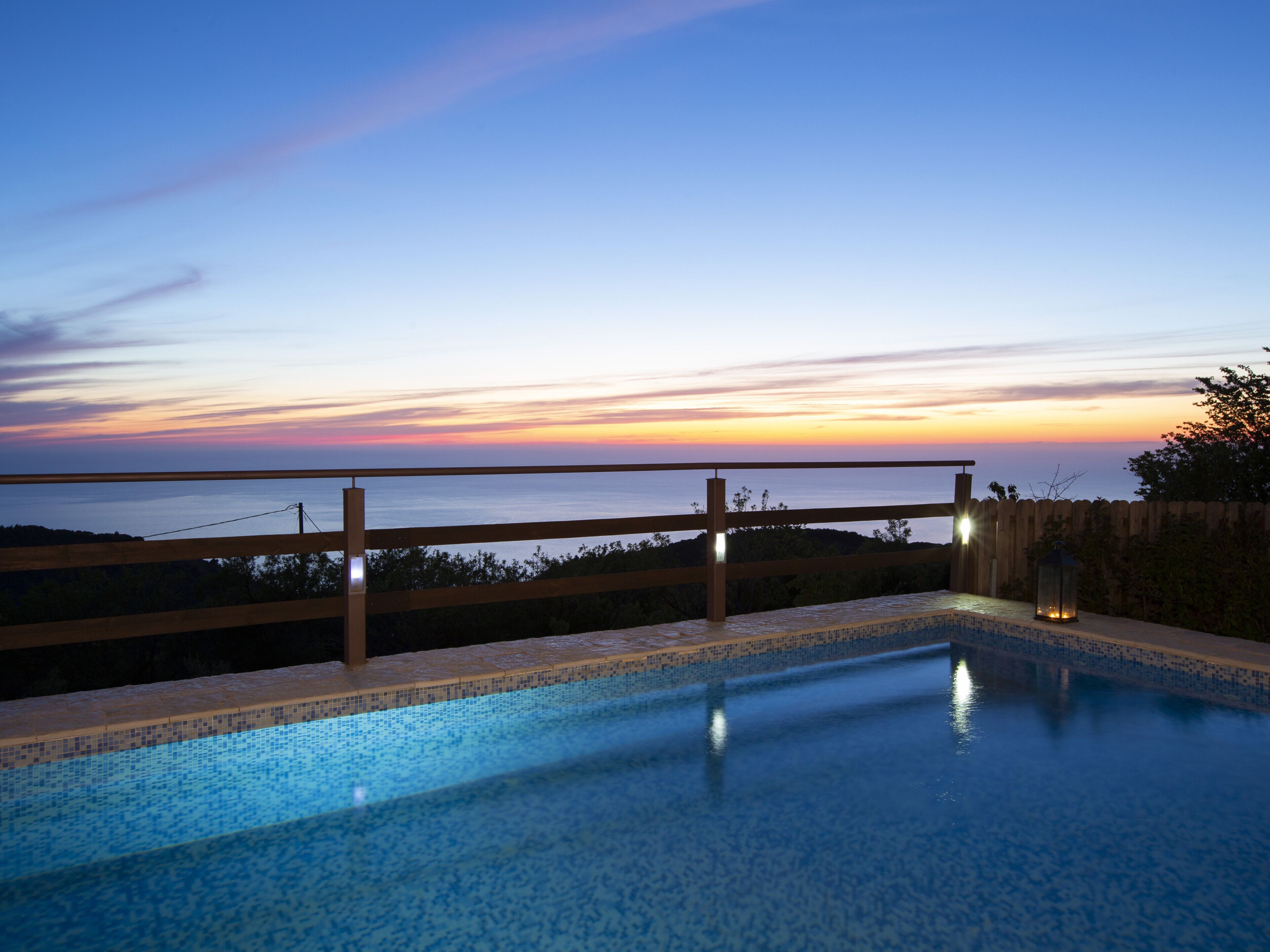 Une piscine dotée d’une terrasse et d’une rambarde en bois, surplombant une étendue d’eau au coucher du soleil.