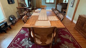 Un comedor con una larga mesa de madera, múltiples sillas, una araña y un tapete floral rojo.