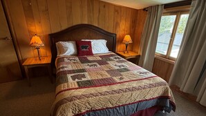 Un dormitorio con pared de paneles de madera, una cama grande con colcha, dos lámparas de noche y una ventana con cortinas.