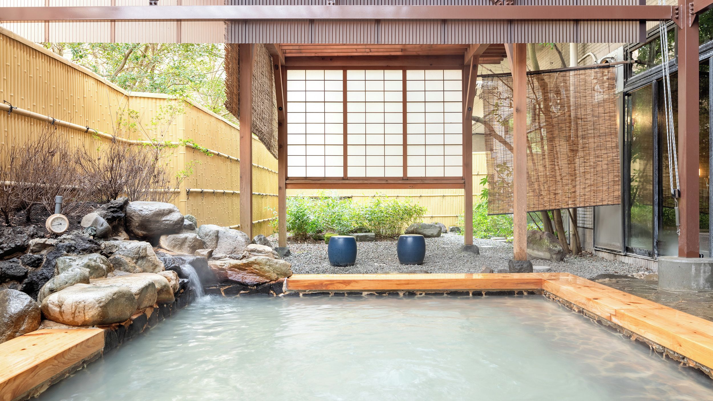 Un onsen japonais traditionnel, doté d’une terrasse en bois, d’un mur de pierre et offrant une vue à travers un paravent en papier.
