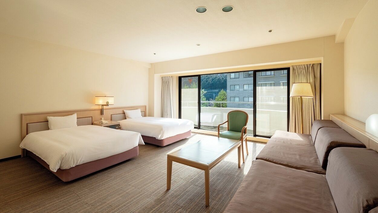 A hotel room with two beds, a sofa, a table, and a view of the outside.