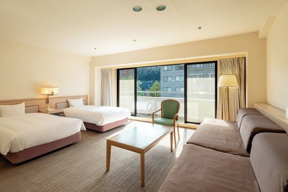 A hotel room with two beds, a sofa, a table, and a view of the outside.