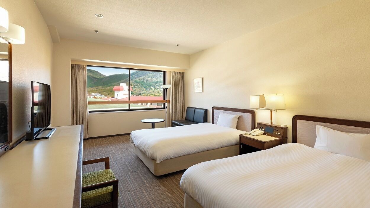 A hotel room with two beds, a desk, a TV, a lamp, and a view of a mountain.