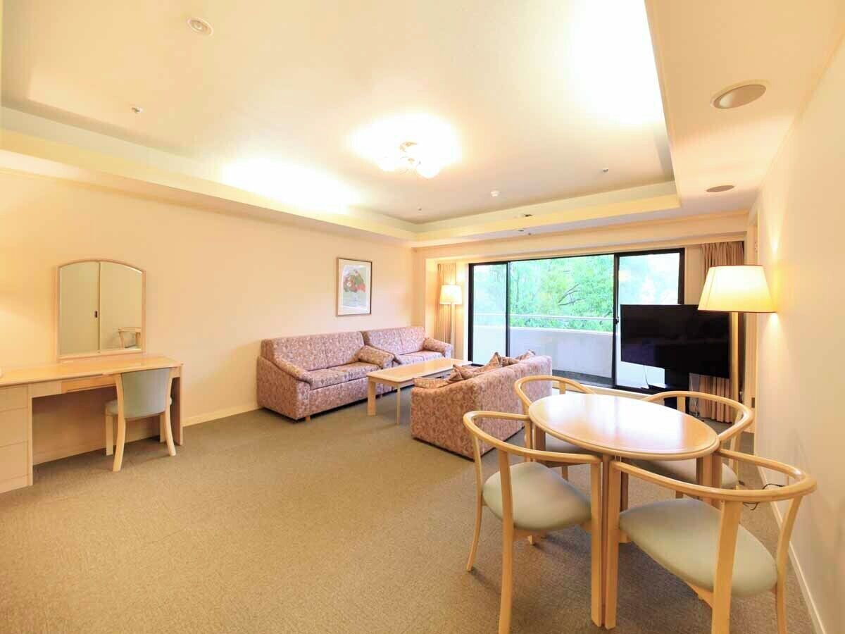 A hotel room with a desk, two sofas, a dining table, a TV, and large windows.