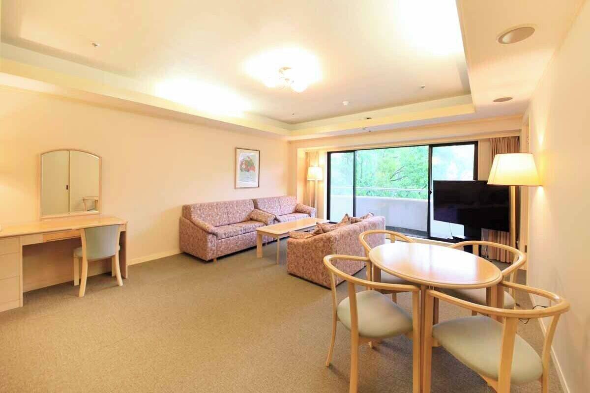 Habitación de hotel con escritorio, dos sofás, una mesa de comedor, un televisor y ventanas amplias.