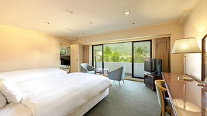Habitación de hotel con una cama grande, un escritorio con televisor, una silla y un balcón con vistas a zonas verdes.