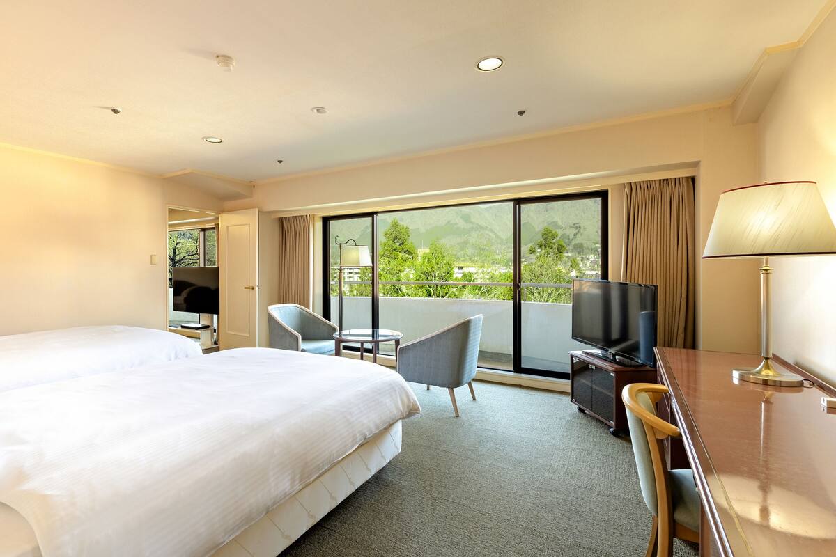 Habitación de hotel con una cama grande, un escritorio con televisor, una silla y un balcón con vistas a zonas verdes.