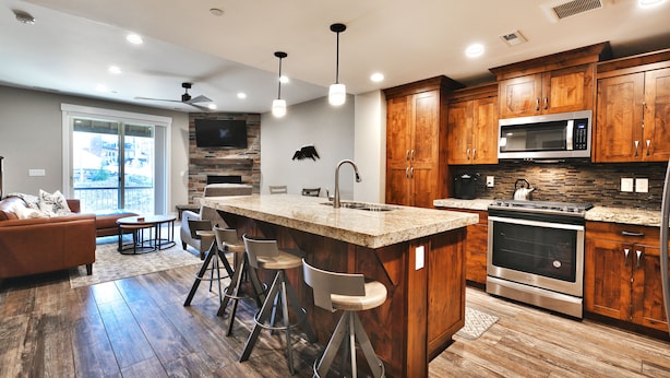 Een moderne keuken met houten kastjes, een centraal eiland met barkrukken, een inbouwoven en een groot raam met uitzicht op een balkon.