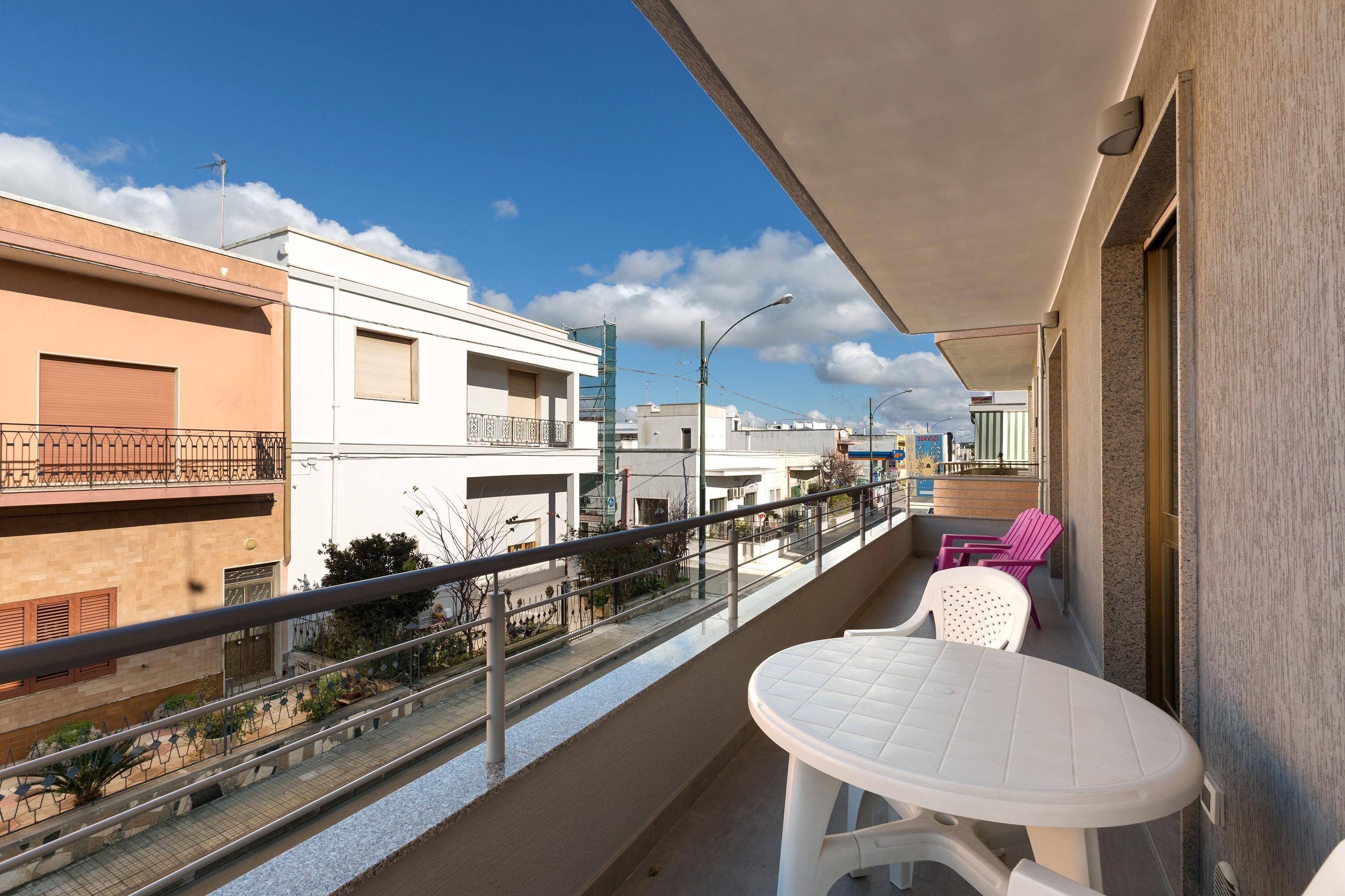 Un balcon avec une table blanche et deux chaises, donnant sur une rue bordée d’immeubles et offrant une vue sur un ciel dégagé.