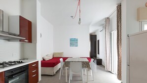 Uma cozinha compacta com fogão, forno e armários, uma área de refeições com mesa e cadeiras, e uma cama com cobertor vermelho.