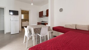 Quarto compacto com cama, mesa de jantar com cadeiras, cozinha compacta, televisão e relógio na parede.