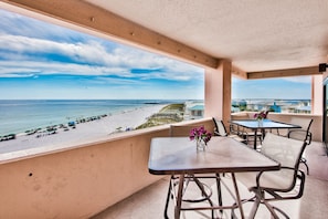 Un balcon avec vue sur une plage et l’océan, agrémenté de tables et de chaises, ainsi que d’un vase rempli de fleurs.