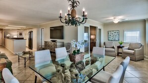 Un amplio salón con una mesa de comedor de vidrio rodeada de sillas, una lámpara moderna y una cocina al fondo.