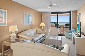 A hotel room with a sofa, armchairs, a coffee table, a TV, and a balcony with a view of the ocean.