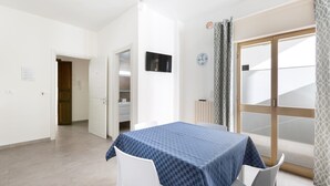Una habitación con mantel de cuadros, sillas blancas, un televisor en la pared y un ventanal corredizo con cortinas estampadas.