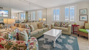 A spacious living room with a large sofa, a coffee table, and a TV. There are multiple windows with blinds, a mirror, and a rug with a sea turtle design. - Bright, designer decorated unit. One free beach chair set up daily. (Destin)