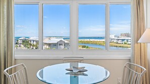A room with a round glass table, two chairs, and a view of a beach and buildings through large windows. - Bright, designer decorated unit. One free beach chair set up daily. (Destin)