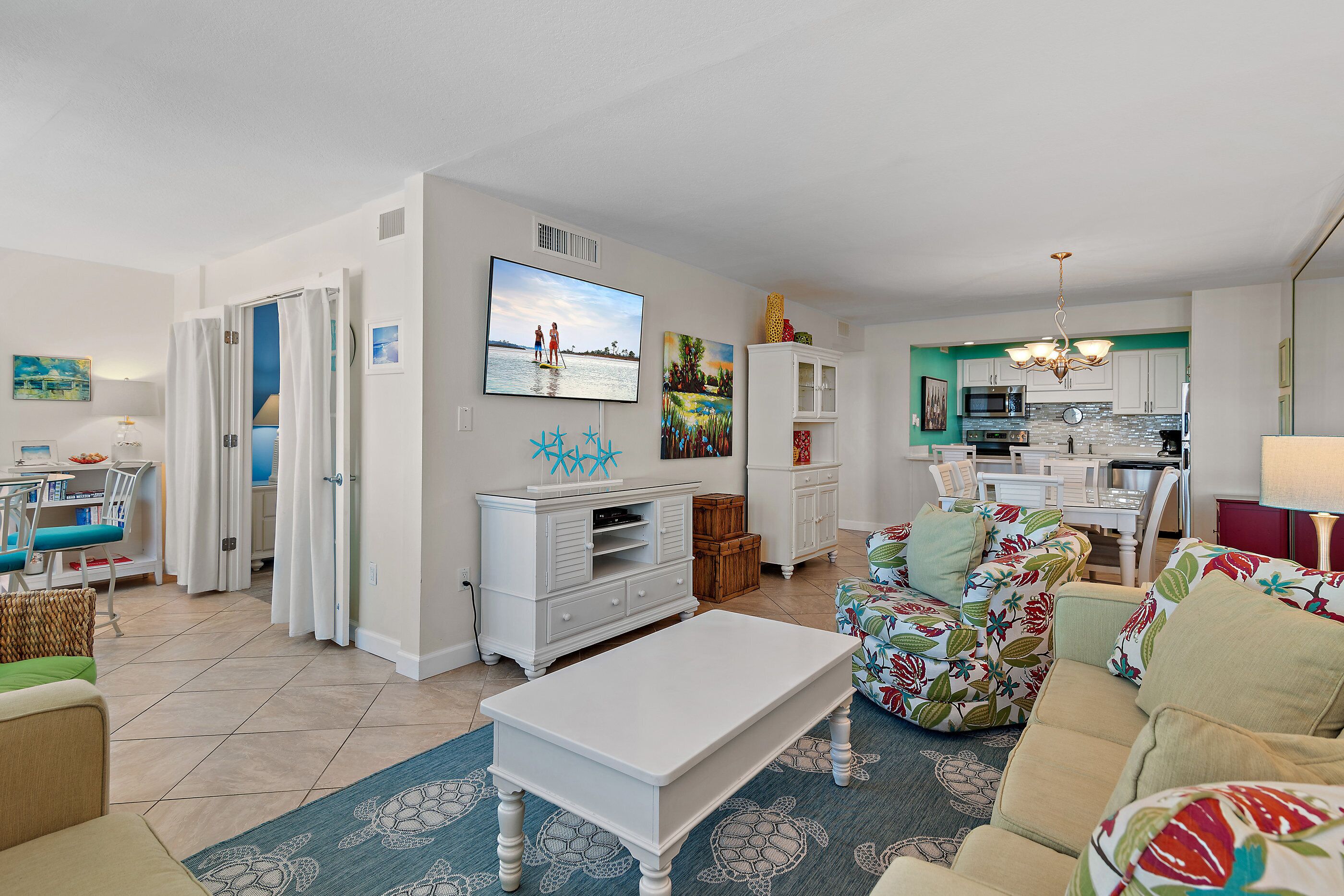 A modern living room with a flat-screen TV, white furniture, and a kitchen area with white cabinets and a microwave.