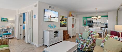A modern living room with a flat-screen TV, white furniture, and a kitchen area with white cabinets and a microwave.