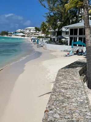 A beach with a stone wall, lounge chairs, and umbrellas.