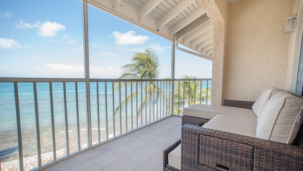 A balcony with a view of the ocean and palm trees, wicker furniture, and a tiled floor.