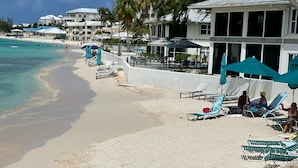 A beachside hotel with sun loungers and umbrellas, a clear view of the ocean, and a white building with large windows.