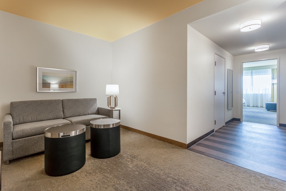 A modern hotel room with a grey sofa, two black side tables, a framed artwork, and a hallway leading to another room.