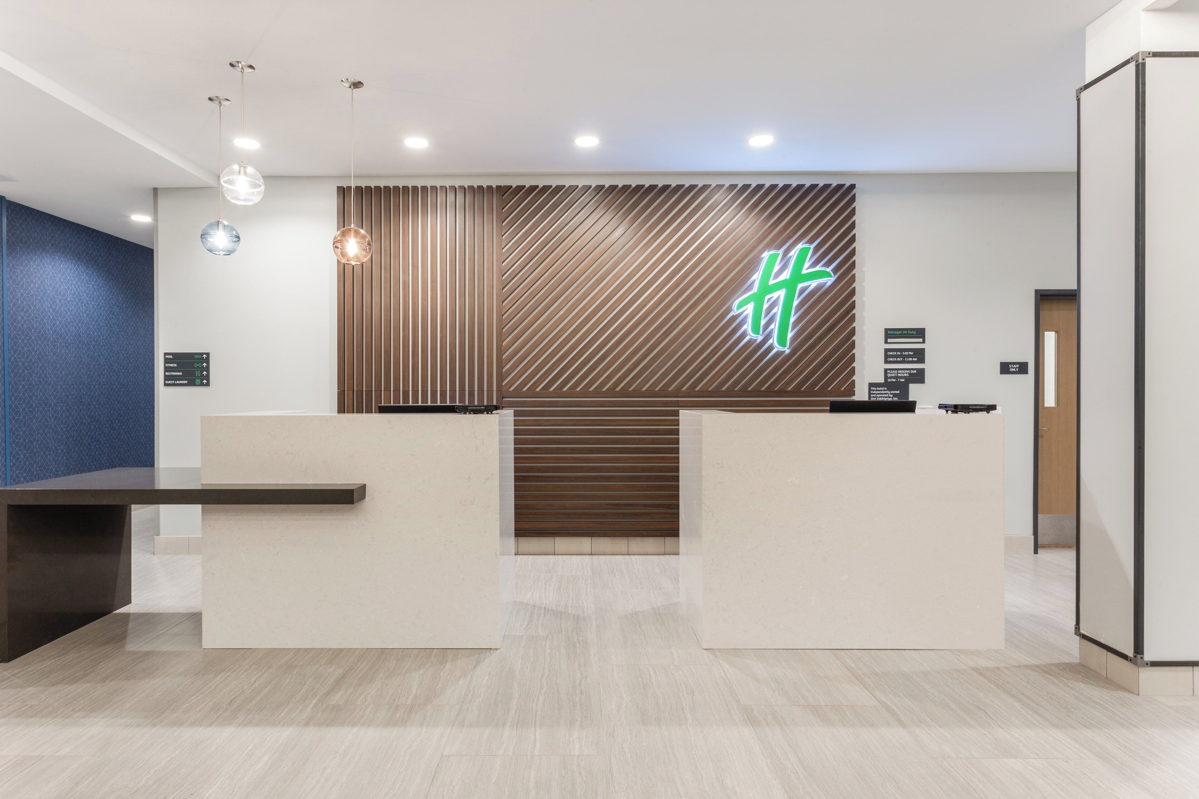 A hotel reception area with a modern design, including a reception desk, a wall-mounted sign with a logo, and directional signs for pool, fitness, and guest laundry.