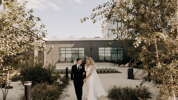 A couple walking through an outdoor area with a modern building in the background. - Holiday Inn & Suites Cedar Falls - Waterloo Event Ctr by IHG (Cedar Falls)