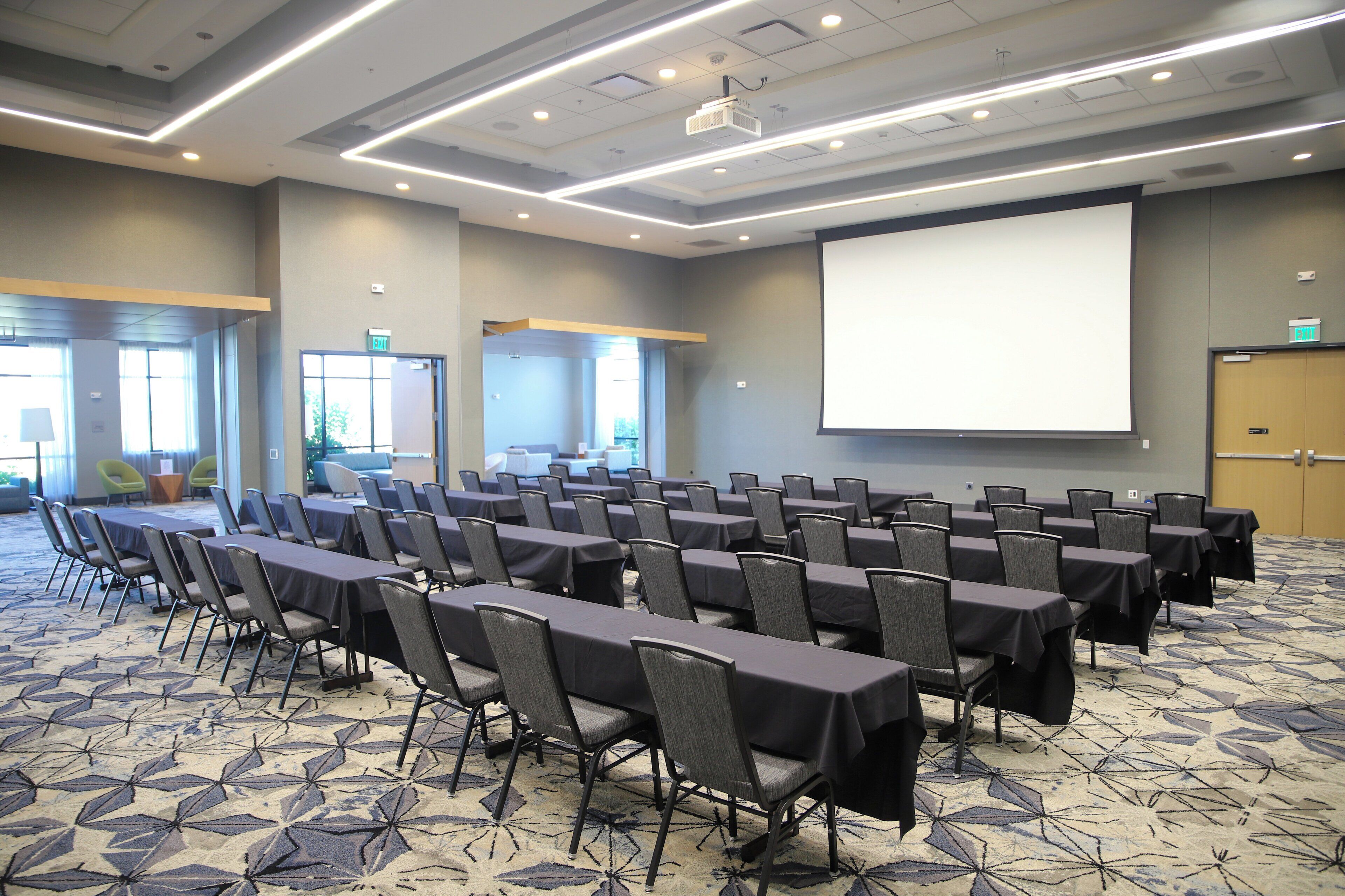 A conference room with rows of tables and chairs, a projector screen, and a carpeted floor.