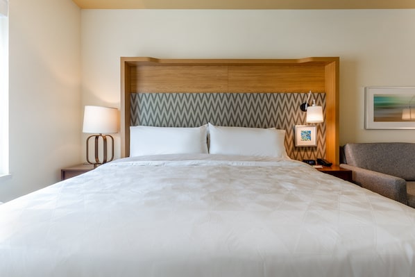 A hotel room with a large bed, a wooden headboard, a nightstand with a lamp, a chair, and a framed picture on the wall.