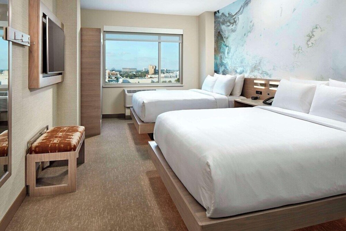 A hotel room with two beds, a TV, a window with a city view, and a painting on the wall.