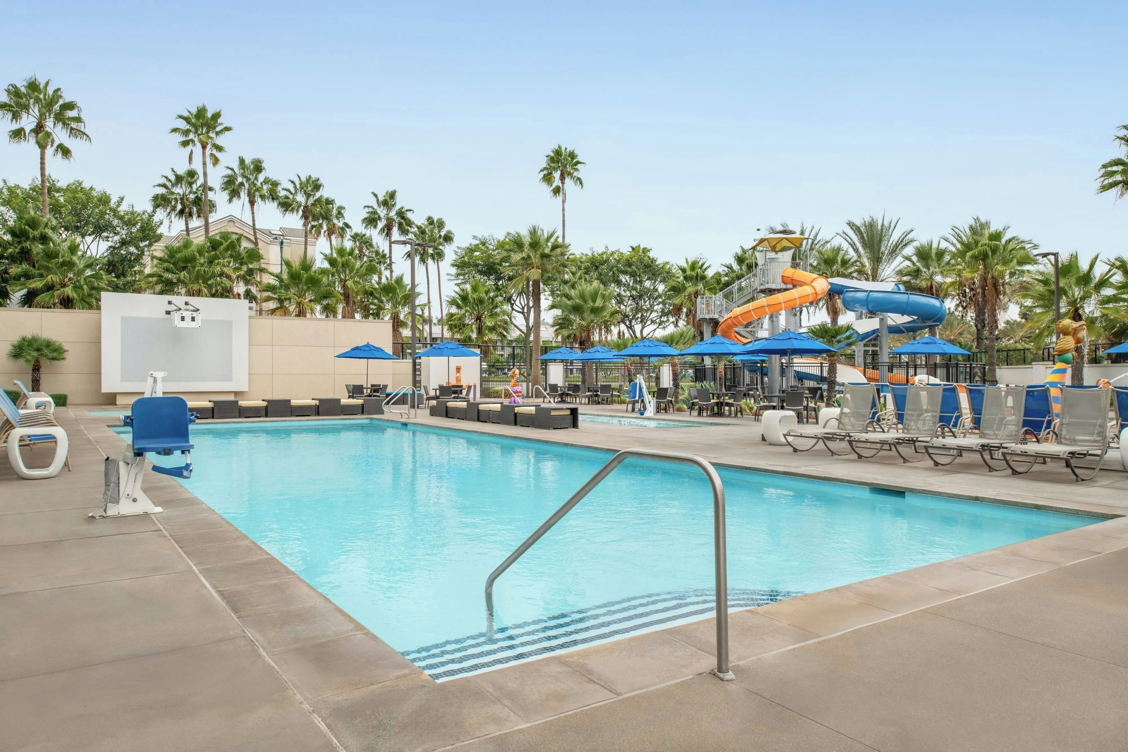 A swimming pool with a water slide, lounge chairs, umbrellas, and palm trees.