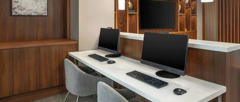 A modern office space with two desks, each with a computer monitor, keyboard, and mouse. There are two grey chairs, a wooden shelf with books and decorative items, and a framed picture on the wall.