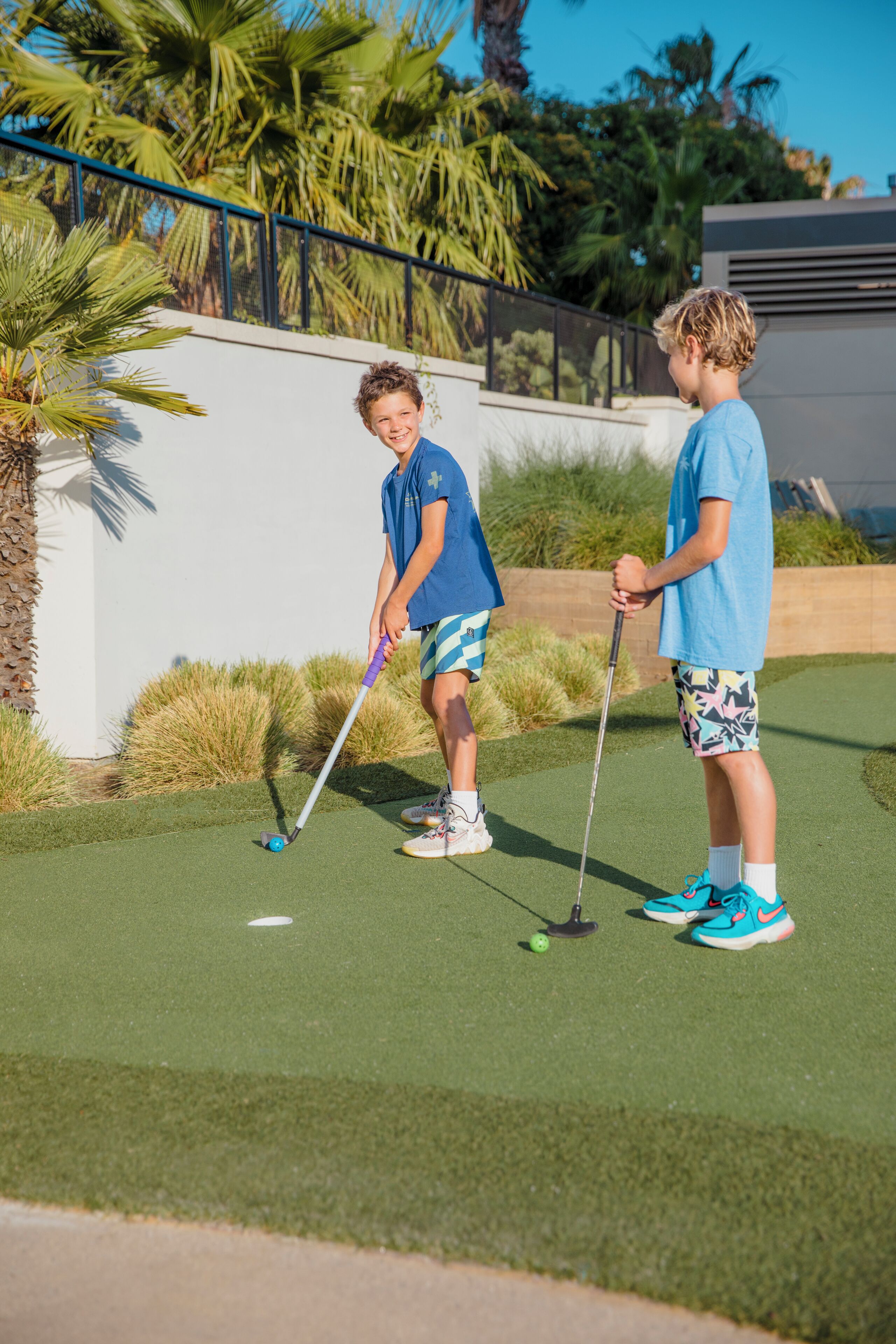 Two people playing golf on a green with palm trees and a modern building in the background.