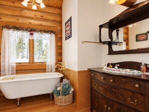 Baño de una cabaña de madera tradicional con una bañera tipo patas de león, un tocador de madera con varios cajones y un espejo grande sobre el lavabo.