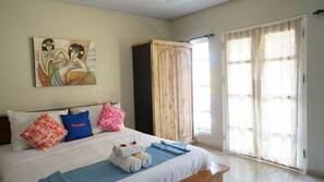 Een net opgemaakt bed met een blauwe deken, witte kussens en een blauw kussen met de tekst 'Matahari'. Aan de muur hangt een schilderij, er staat een houten garderobe en er is een groot raam met gordijnen.