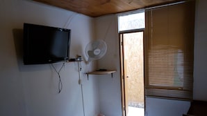 Un dormitorio con cama, televisor montado en la pared, ventilador de pie y ventana con persianas.