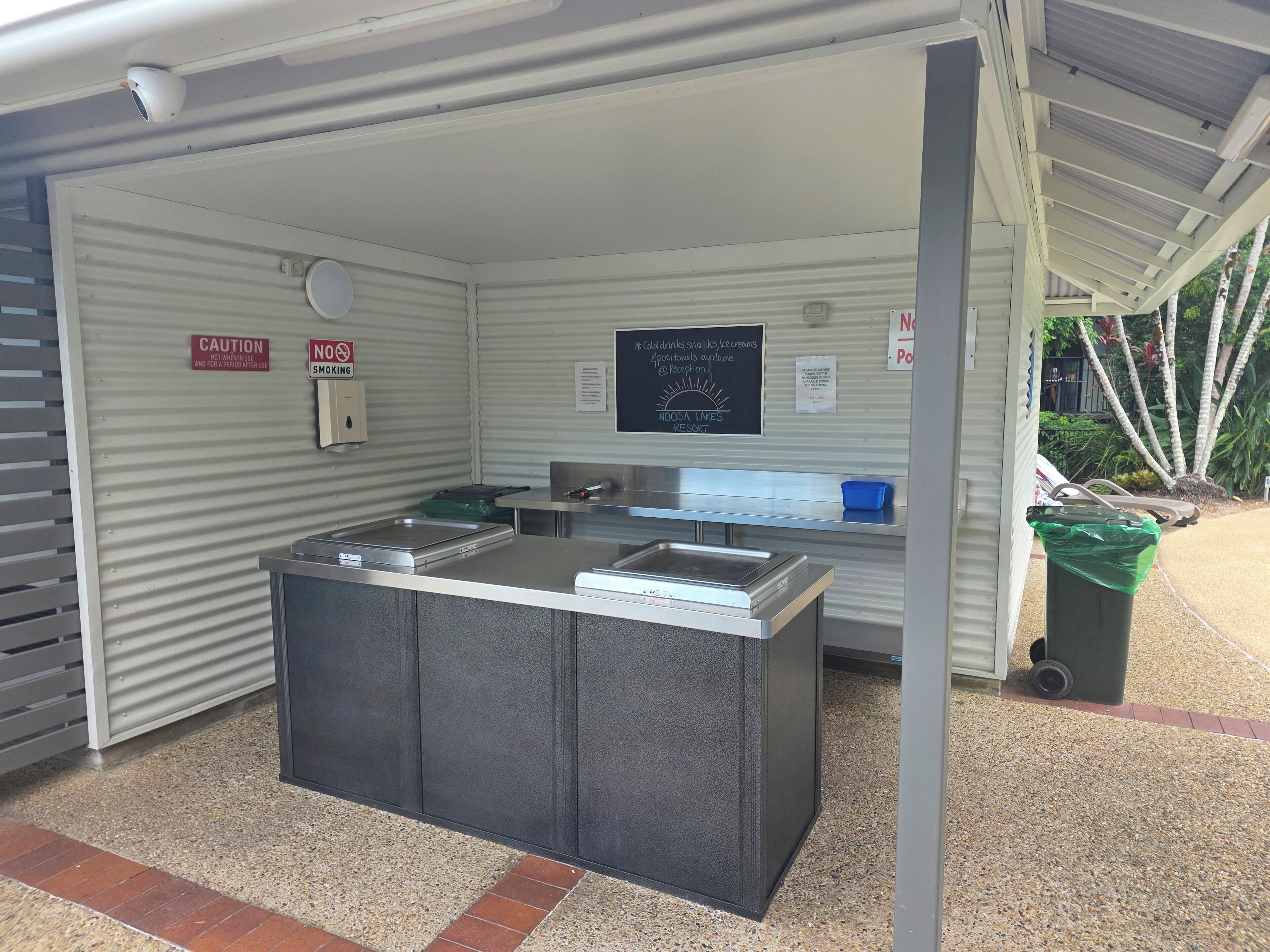 A self-service beverage station with a caution sign, a no smoking sign, and a sign for Noosa Lakes Resort.