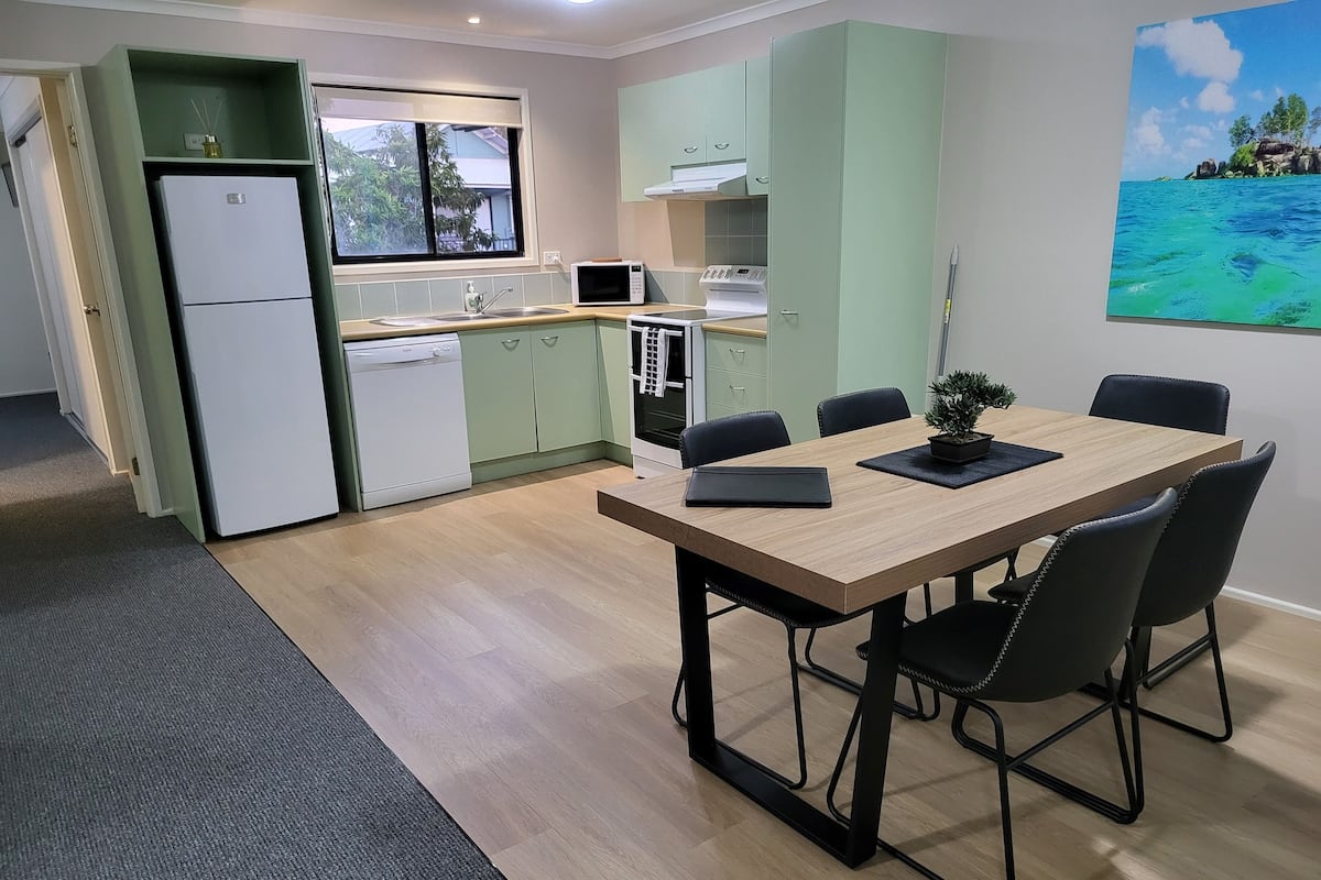 Een compacte keuken met een vaatwasser, magnetron en kookplaat. Er is een zithoek met een tafel en stoelen, en een schilderij van een strandzicht aan de muur.