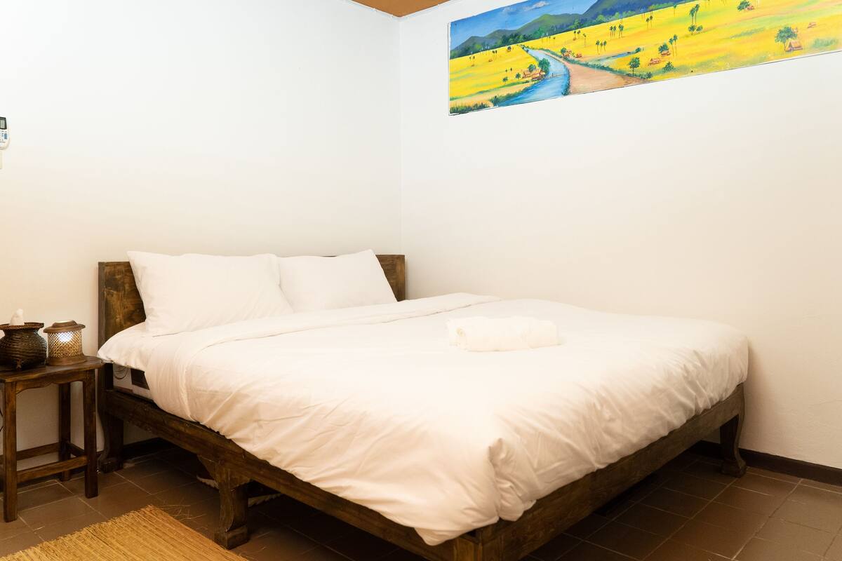 A bedroom with a wooden bed, white bedding, a small bedside table with a lamp, and a painting of a landscape on the wall.