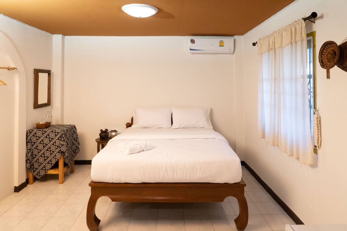 A bedroom with a wooden bed, a bedside table with a patterned tablecloth, a hanging basket, and a wall-mounted air conditioner.