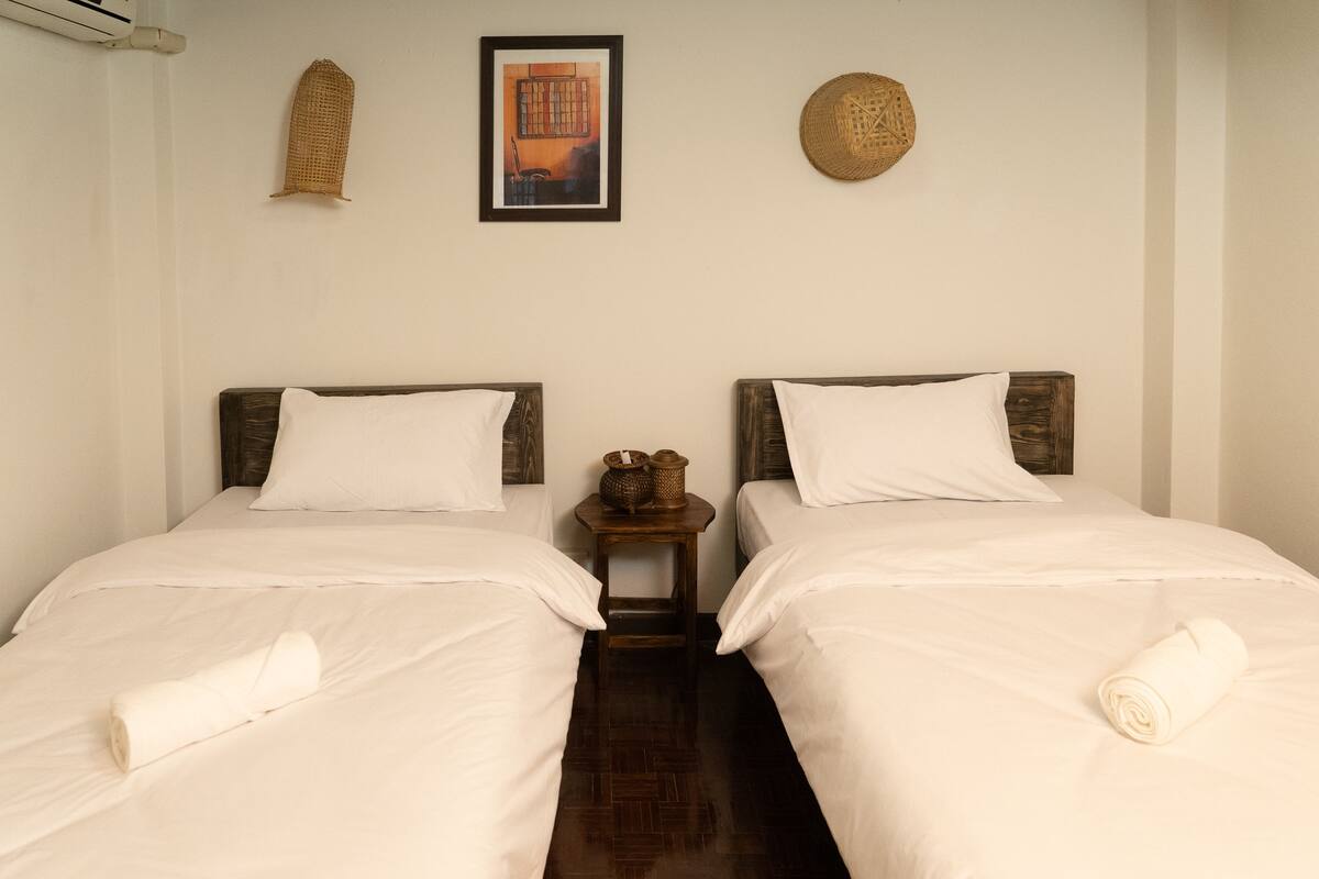 A double room with two beds, a small wooden nightstand, a woven wall hanging, and a framed picture.